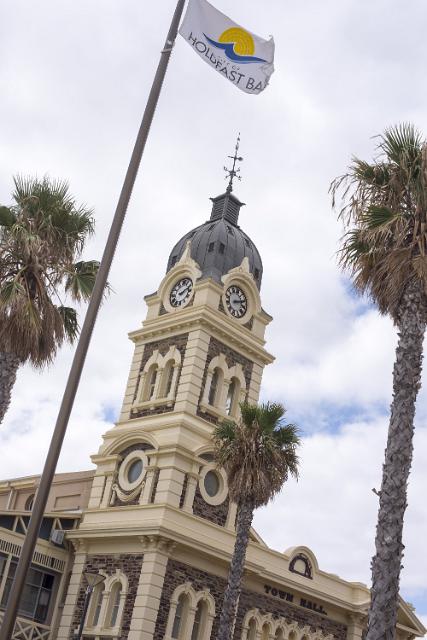 The historic clock tower on the town hall in Glenelg, a suburb of Adelaide, South Australia flanked by tropical palm trees and a fluttering flag