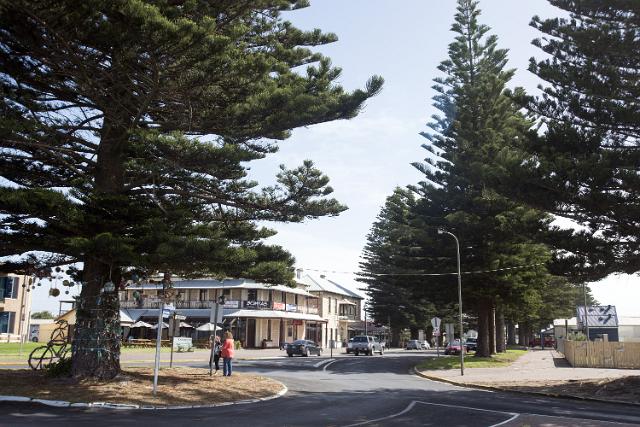 Street scene with people and bicycles under tall pine trees in front of an old heritage building, Beachport, Australia